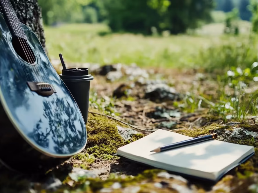 Acoustic guitar leaning against a tree with a notebook and pencil on mossy ground, symbolizing a peaceful outdoor songwriting camp setting