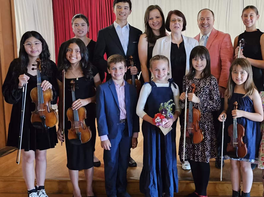 K&M Music School students and teachers holding violins smile together on stage after a community recital, illustrating accessible music education.